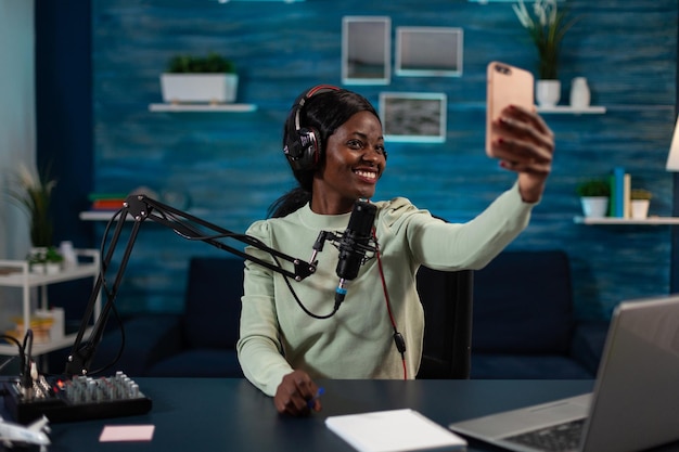 Woman vlogger holding smartphone smiling at camera while taking photo during entertainment vlog. Social media creator recording podcast answering subscribers questions using production equipment