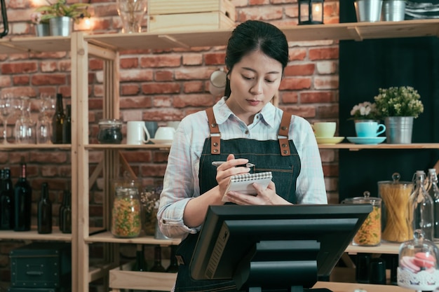 カウンターに立って喫茶店で客から注文を受けている女性ウェイトレス。メモに書き込み、デジタルタブレットを使用して喫茶店でアジアの日本人女性労働者。 posマシン端末の概念。