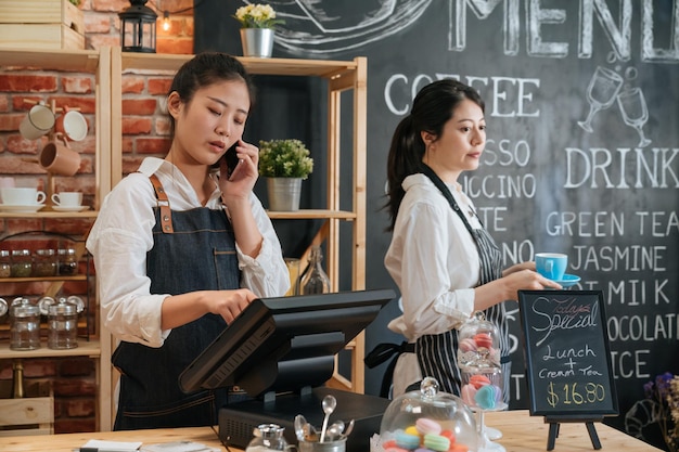 女性のウェイトレスは、デジタルタブレットで作業し、スマートフォンで話し、現代のコーヒーショップで注文を受け付けています。中小企業のオンライン購入とサービスの概念。同僚のバリスタが歩いて食事を送る