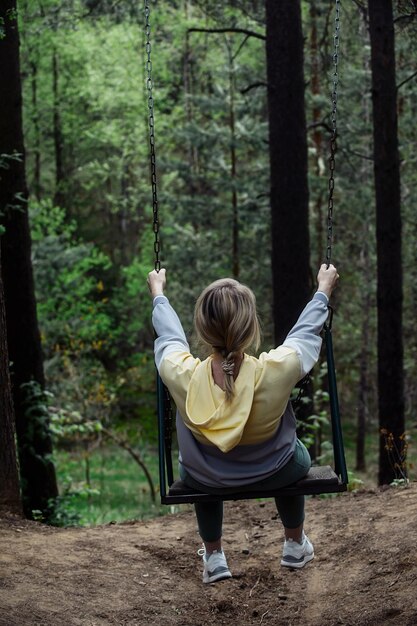 a woman on a walk in a green forest rides on a high swing attached to tall trees