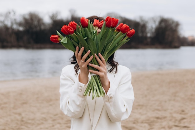 Woman walking on the beach shore in moody cloudy windy weather with bouquet of red tulips flowers