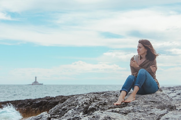 Woman walking by rocky sea beach in wet jeans lighthouse on background