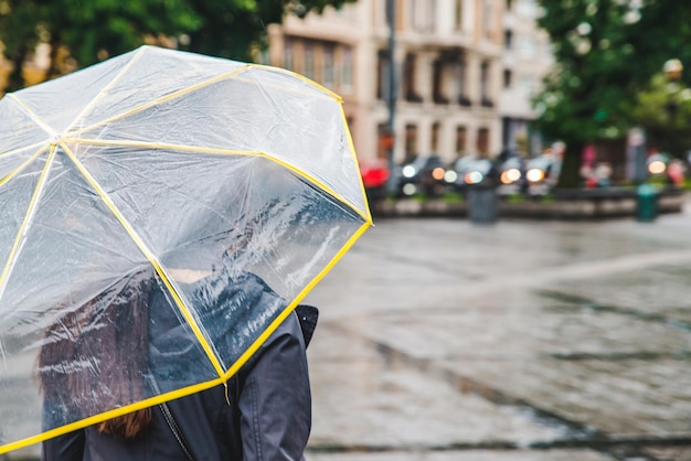 Woman walking by wet rainy streets under transparent umbrella copy space