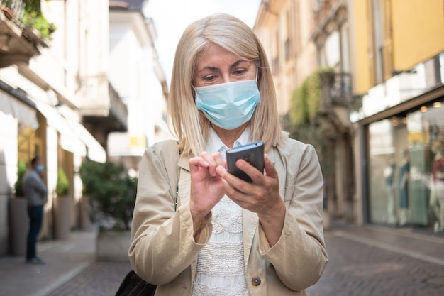 Фото Женщина гуляя на улицу города пока использующ ее smartphone во время пандемии коронавируса