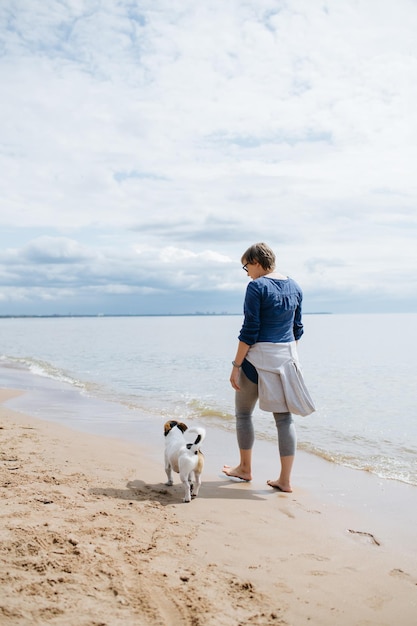 모래 해변에서 그녀의 강아지와 함께 산책하는 여자 후면보기