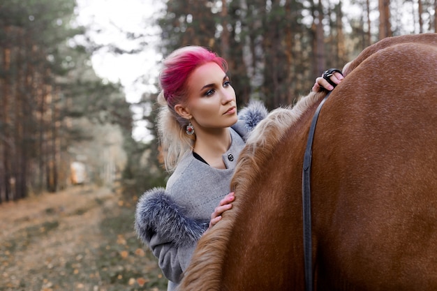 Woman walking with horse autumn on nature. Creative hot pink makeup girl face, hair coloring. Portrait of a girl with a horse. Horse riding in the autumn forest. Autumn clothes and bright red makeup