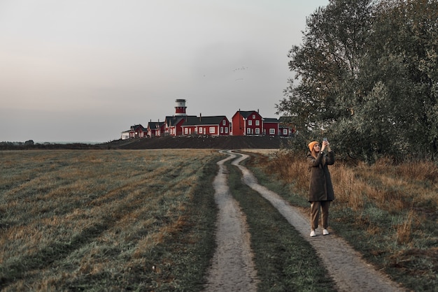 A woman walks along a country road, a red and white lighthouse at the end of the road. Travel, dawn.