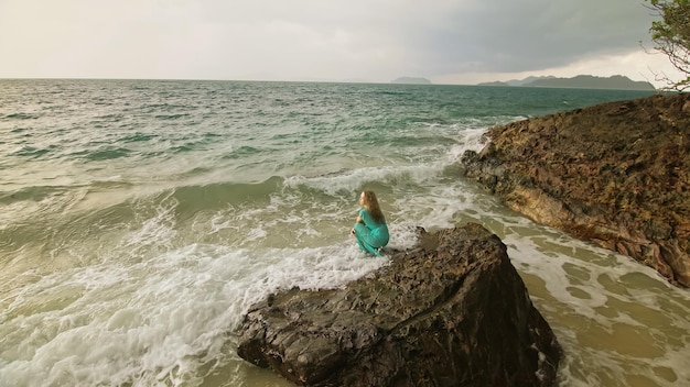 여자는 바다 암초 돌 폭풍우 흐린 바다 파란색 수영복 드레스 튜닉 개념 휴식 열대 리조트 해안선 관광 여름 휴가의 바위에 산책