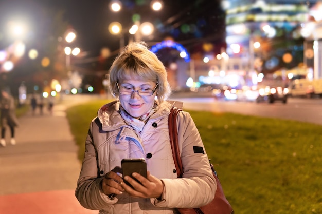 A woman walks through the city at night. A middle-aged woman with a phone in her hands is on the sidewalk.