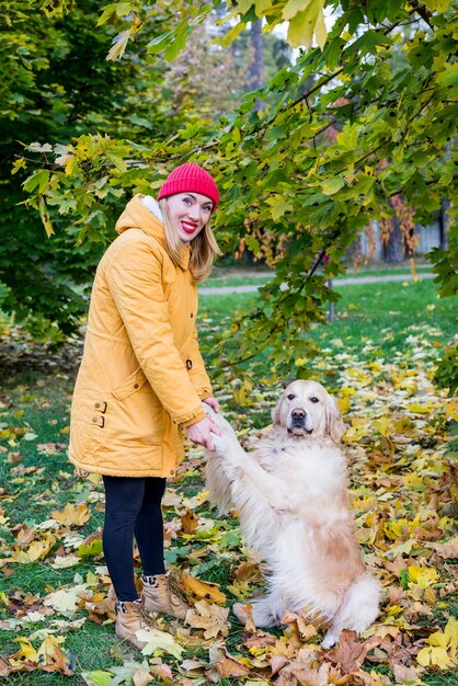 暖かい服を着た女性は、秋の黄色の葉の間で前足で彼女のレトリバーを保持します