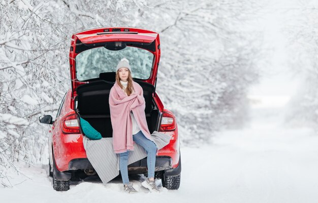 Женщина в теплой одежде сидит в зимнем лесу, опираясь на машину и держа чашку кофе.