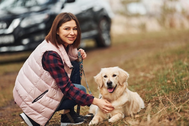 現代の黒い車に対して森の中で彼女の犬と一緒に座っている暖かい服を着た女性