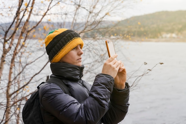 A woman in a warm jacket photographs nature in late autumn dressed in a yellow hat takes pictures of nature and forests by the river or lake outdoor