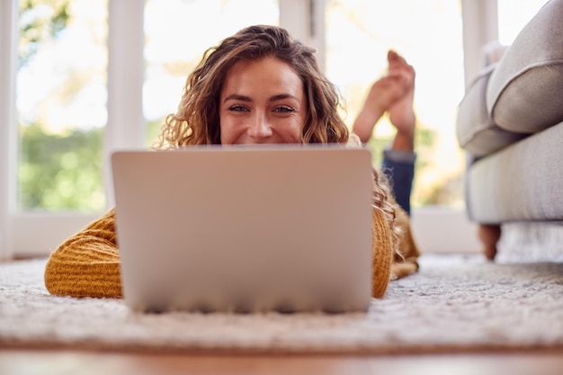 Woman In Warm Jumper Lying On Floor At Home Using Laptop