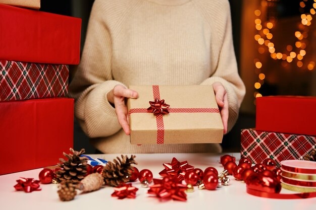 Woman in warm sweater holding gift box wrapping in craft paper with festive ribbon present for winte