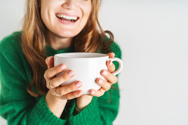 Photo woman in warm wool green sweater is holding white mug in her hands with coffee or tea. mock up for winter mood design. minimal style.