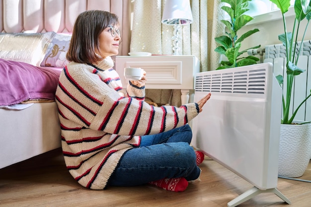 woman warming in bedroom near electric heater middleaged female in woolen sweater resting at home near heating radiator with cup of coffee in hands lifestyle cold heating season autumn winter