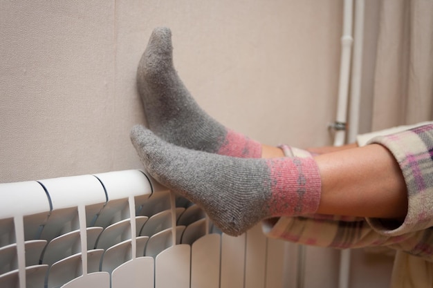 a woman warms her feet on a heating radiator Heating concept in an apartment