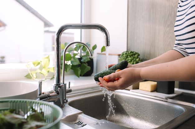 Donna che lava i cetrioli verdi per insalata in cucina nel lavandino sotto l'acqua corrente.