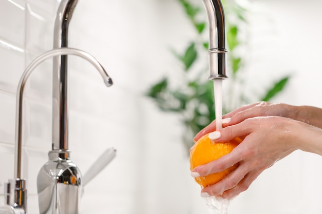 シンクキッチンの蛇口の下で熟したオレンジを洗う女。