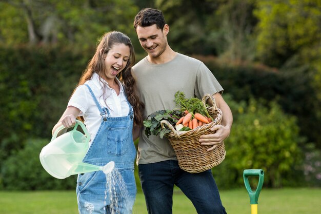 Woman watering a plants while man holding basket of vegetables in garden