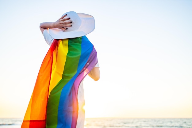 Woman waving with rainbow flag and hat at sea beach at sunset