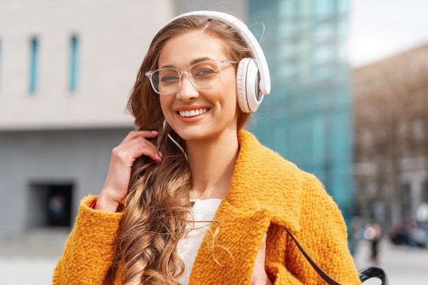 Woman wear eyeglasses listen music headphone outdoor walking outdoor city street dressed stylish yellow coat close up portrait smile Caucasian female 30s enjoy podcast or audio books outside