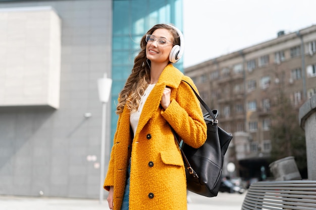 女性は眼鏡をかけます音楽を聴きますヘッドフォンウォーキング屋外シティストリート服を着たスタイリッシュな黄色のコートと黒のバックパック笑顔白人女性30代外でポッドキャストやオーディオブックをお楽しみください