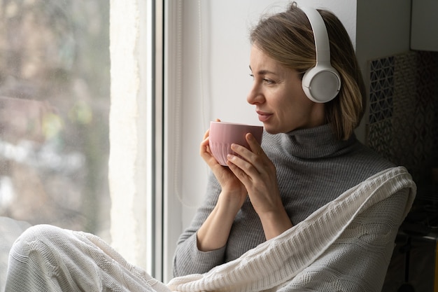 Woman wear wireless headphones, listening to music, sitting on the windowsill, looking at window