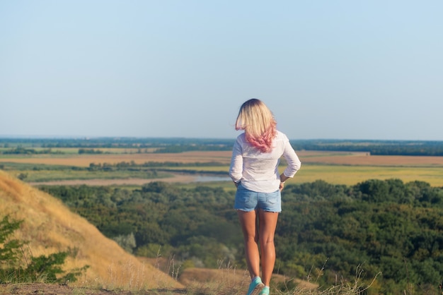 woman weared white shirt and blue shorts with long blonde pink hair standing on the mountain