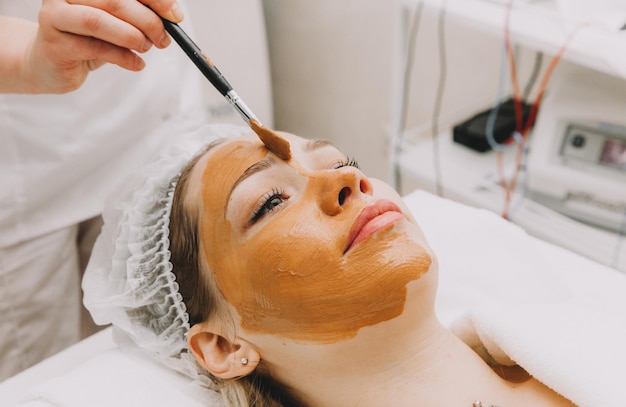 Фото Женщина в маске на лице в салоне красоты косметолог наносит глиняную маску на лицо девушки