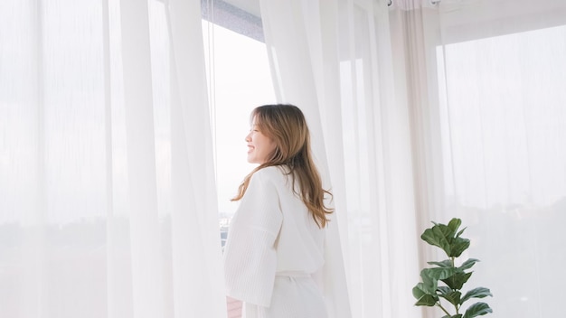 Woman wearing bathrobe opens curtains in bedroom and see around the outdoors Welcome new sunny day happy positive young woman wakes up and looks in the window at home or hotel feeling optimistic