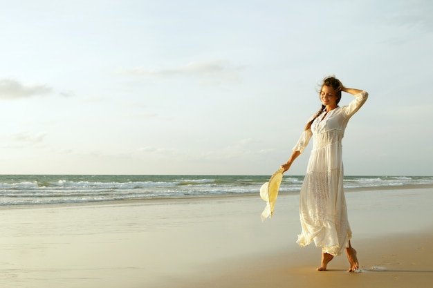 Woman wearing beautiful white dress is walking on the beach during sunset