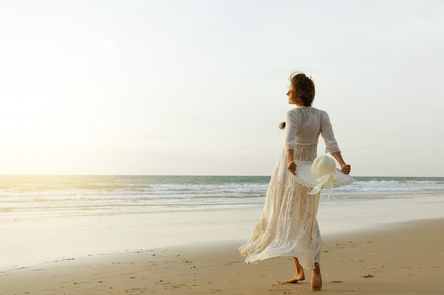 Woman wearing beautiful white dress is walking on the beach during sunset