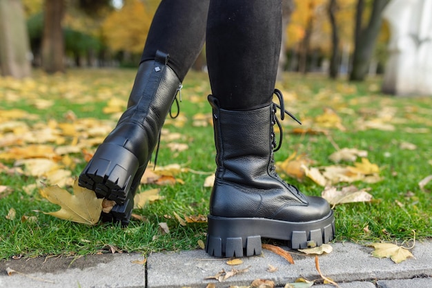 A woman wearing black boots and black boots stands on a curb in a park.