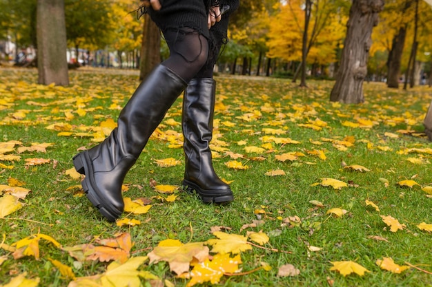 A woman wearing black boots is walking in the grass with yellow leaves on the ground.