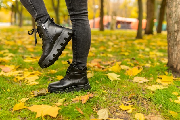 A woman wearing black boots stands in a park with yellow leaves on the ground.