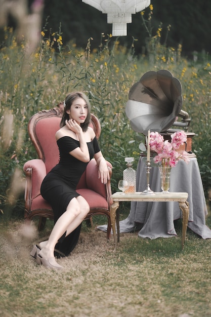 Woman wearing black dress sitting on the couch on flowers garden with vintage gramophone and decoration.