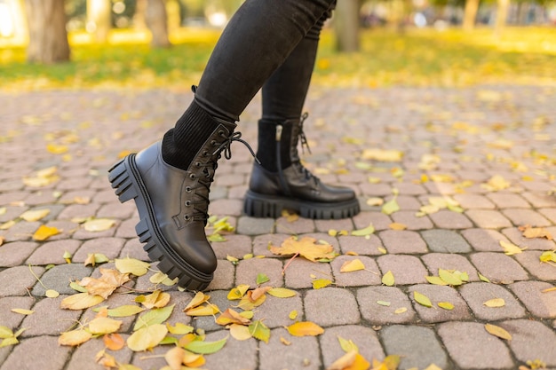 A woman wearing black leather boots stands on a brick walkway in autumn leaves.