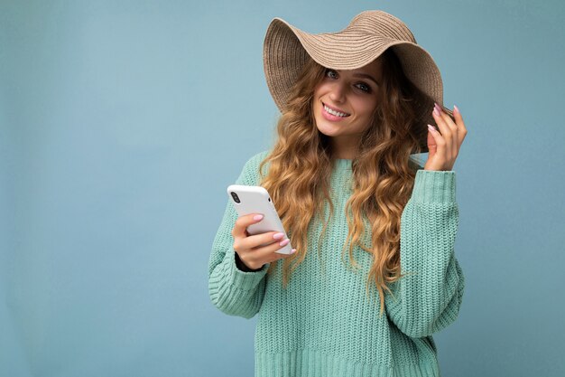 青いセーターと帽子を身に着けている女性は、カメラを見て電話を介してインターネット上でサーフィンの青い背景の上に孤立して立っています。