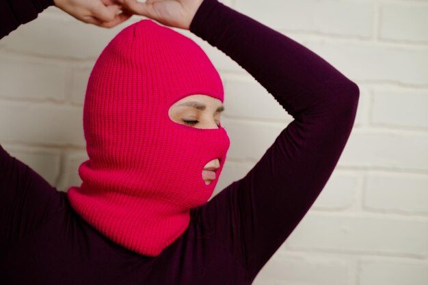 a woman wearing a bright pink balaclava dances energetically striking unique poses with her hands against a clean white brick wall background