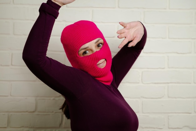 a woman wearing a bright pink balaclava dances energetically striking unique poses with her hands against a clean white brick wall background