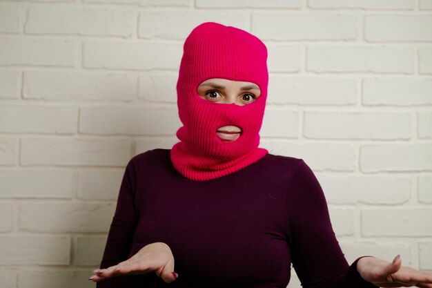a woman wearing a bright pink balaclava dances energetically striking unique poses with her hands against a clean white brick wall background