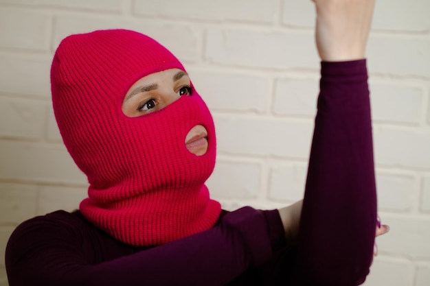 a woman wearing a bright pink balaclava dances energetically striking unique poses with her hands against a clean white brick wall background
