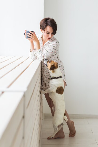 Woman wearing cozy home shirt relaxing at home and playing with dog jack Russell terrier, drinking