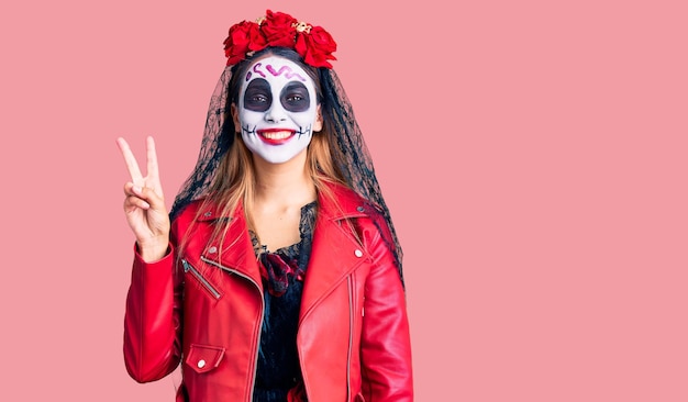 Woman wearing day of the dead costume over background showing and pointing up with fingers number two while smiling confident and happy