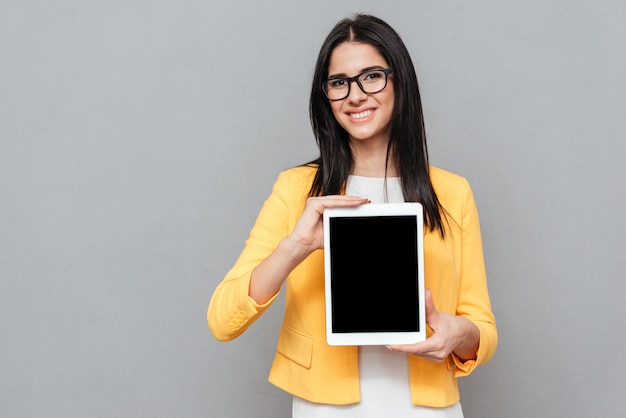 Woman wearing eyeglasses and dressed in yellow jacket showing tablet computer display to front over grey surface. Look at front.