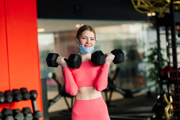 La donna che indossa la maschera per il viso esercita l'allenamento in palestra durante la pandemia del virus corona.