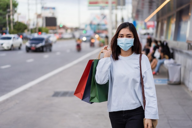 Woman wearing face mask holding smartphone and shopping bag waiting for bus at bus stop in city street