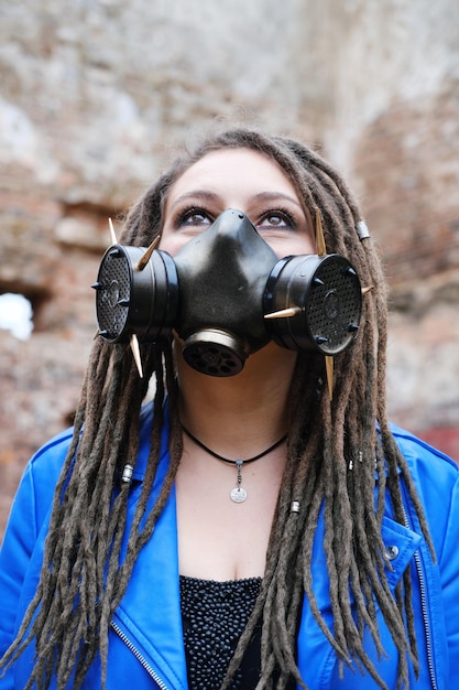 Woman wearing gas mask with spikes in blue jacket and black dress standing in ruined building